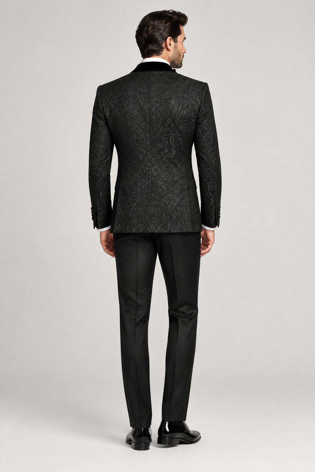 Back view of a model wearing a black patterned tuxedo jacket with matching black trousers.