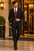 Model wearing a black three-piece suit with a burgundy vest and striped tie at an elegant hotel entrance.